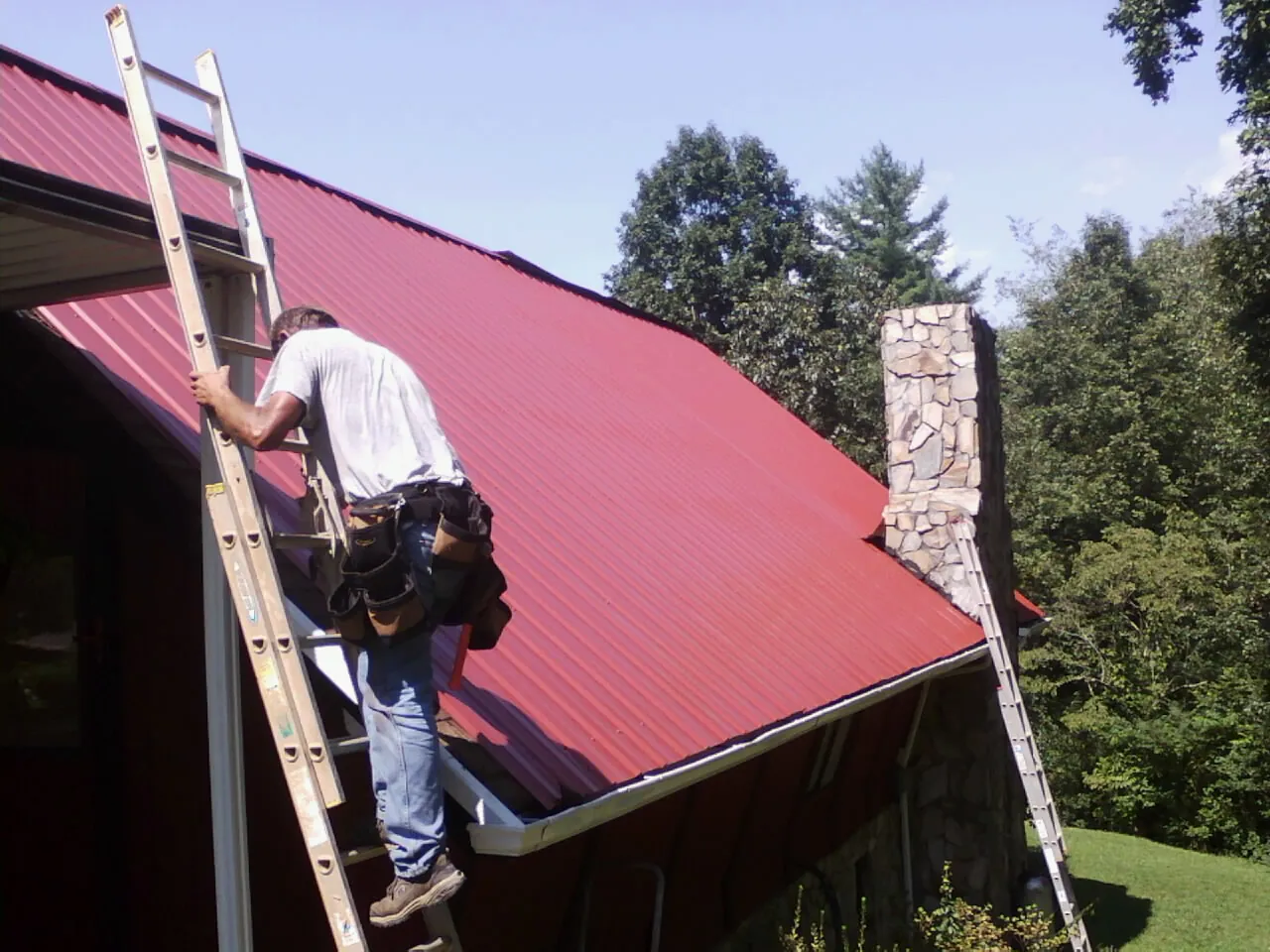 Roofer on a ladder beside a red metal roof and stone chimney — Soffit & Fascia Repair by Roofing Brothers in Colstrip